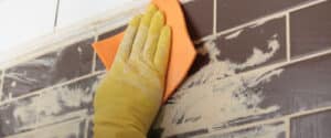 Immagine ravvicinata di una mano con guanto che pulisce con una spugna delle mattonelle marroni sporche di vernice o altro materiale. 