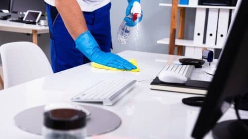 Immagine ravvicinata di un operatore che pulisce con straccio e prodotto una scrivania bianca. Sopra c'è un monitor, una tastiera e un mouse. Sullo sfondo si vedono degli scaffali con dei faldoni.